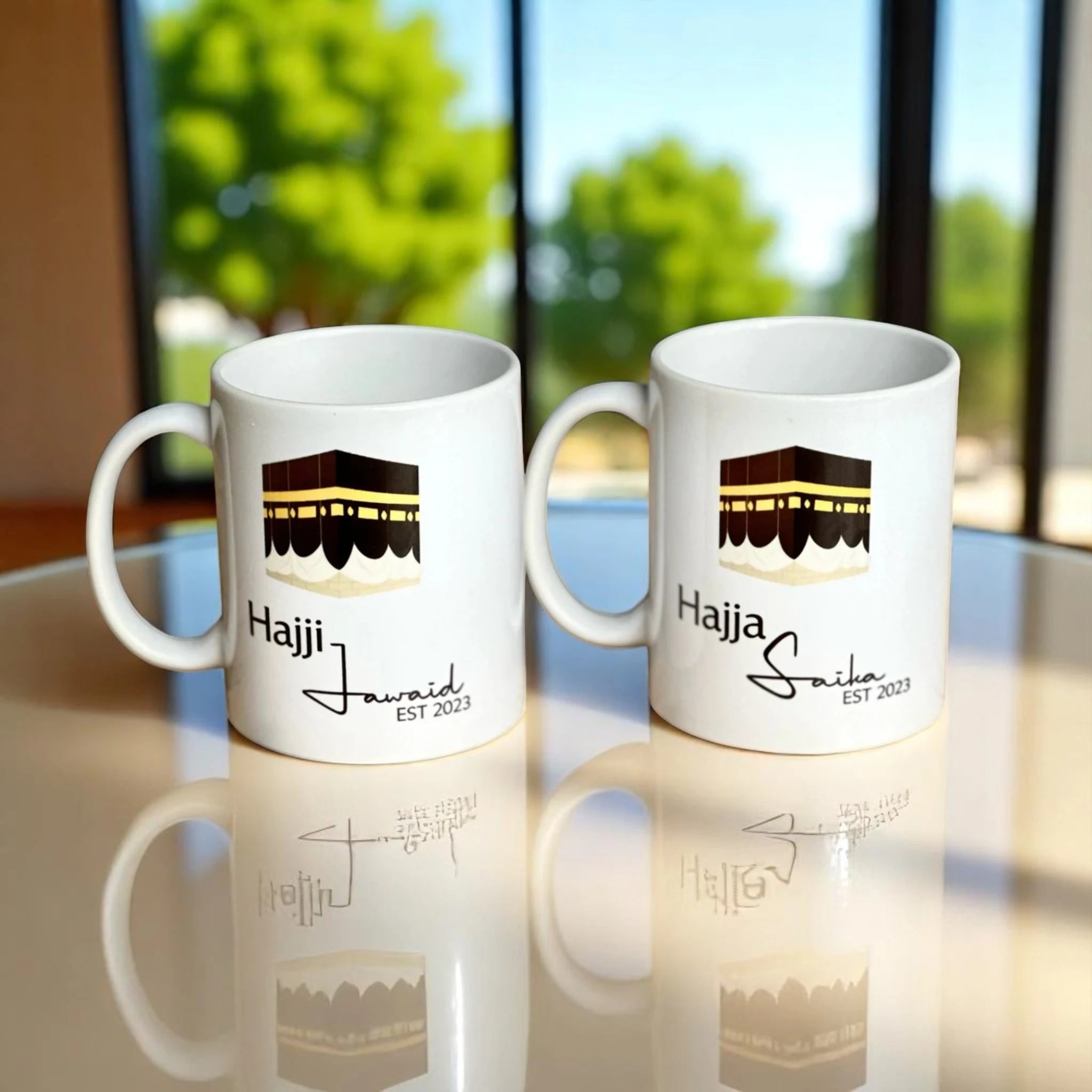 Two white ceramic mugs with 'Hajji' and 'Hajja' titles alongside silhouettes of the Kaaba, placed on a reflective surface with a bright window in the background.