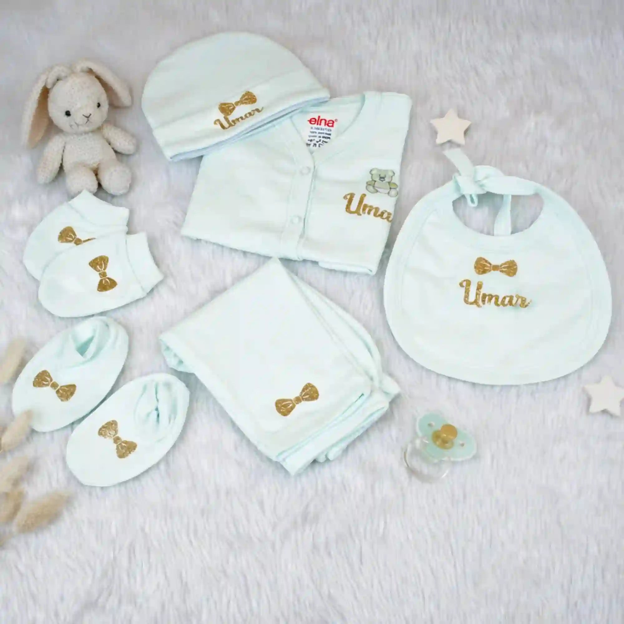 Collection of baby clothing and accessories with 'Umar' embroidered on them, including a onesie, bib, hat, and blanket, on a soft surface.