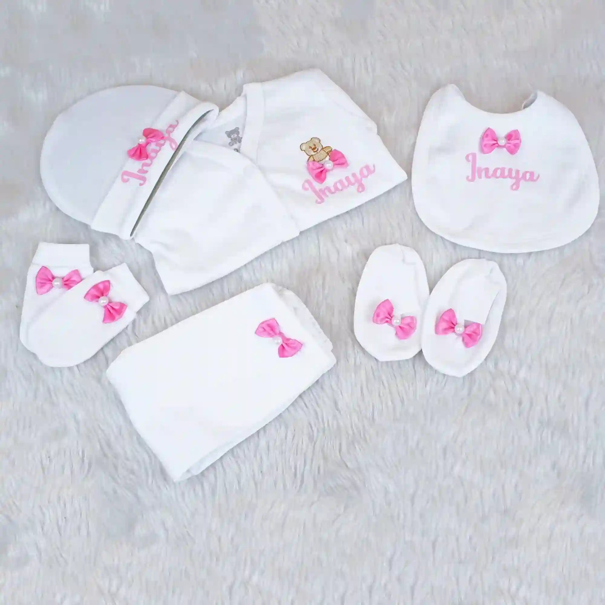Baby clothing set with white bibs, hats, and socks featuring pink bows and embroidered names on a light gray background.