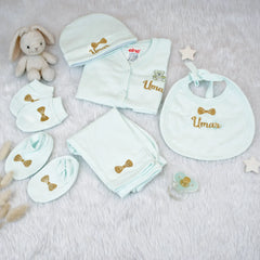 Collection of baby clothing and accessories with 'Umar' embroidered on them, including a onesie, bib, hat, and blanket, on a soft surface.