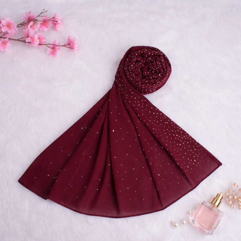 Rolled deep red studded Hijab laid on a soft surface with perfume and flowers, emphasizing its luxurious and gift-worthy appeal.