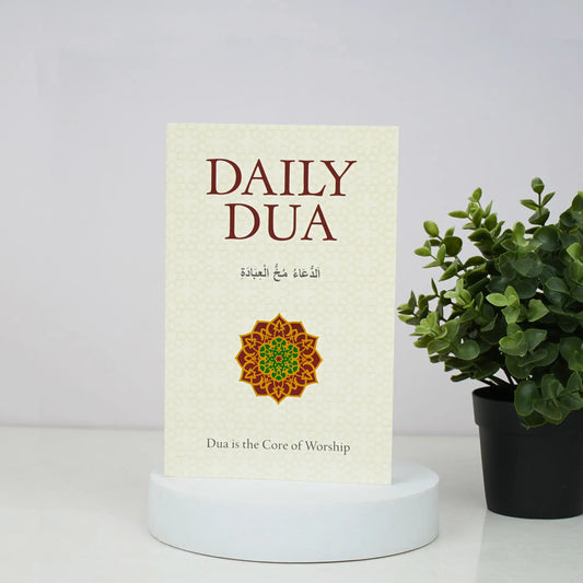 Book titled 'Daily Dua' with a decorative cover on a white surface next to a potted plant.