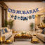 EID Mubarak Luxe Banner hanging across a modern, warmly lit living room decorated for festive Eid celebrations with candles and floral arrangements.