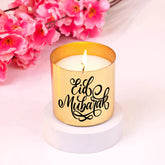 Eid Mubarak Scented Candle in a gold jar with elegant calligraphy, placed on a white base with pink flowers. Luxurious Eid fragrance gift.
