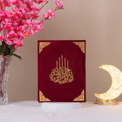 Velvet Quran with gold calligraphy from the Maroon Salah Combo, displayed upright beside crescent lamp and pink flowers for ceremonial appeal.