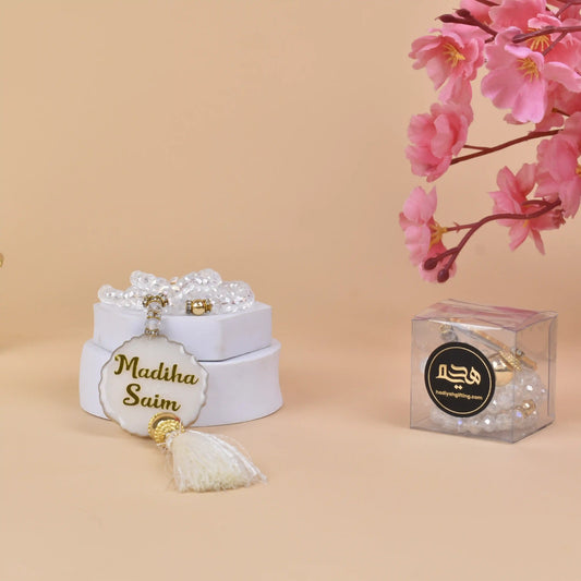 White Crystal Tasbeeh with 100 beads and custom name 'Madiha Saim', displayed in a gift box with floral decor. Ideal Islamic gift for Hajj & Umrah.