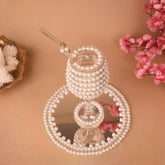 Decorative item with pearls on a reflective surface with flowers and a decorative plate in the background.