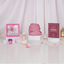 Front display of a pink Graceful Hijab Set women's hamper resting on white display blocks over a glossy surface scattered with pearls and pink petals. The set features a rolled pink hijab, a dark red "Fortress of the Muslim" dua book, a framed pink digital tasbeeh, a pink pearl tasbeeh with a tassel, and a matching tally counter, set against white curtains, fairy lights, and a golden lantern.