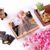 A wooden crate gift basket, wrapped in pink netting with floral and gold ribbon accents, holds a white mug and black tea packets. Surrounding the basket are loose OLITIA tea bags, a glass of tea, a small wooden tray with dried flowers, and a large branch of artificial pink cherry blossoms on a white surface.