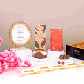 A gift hamper setup with a customised round couple frame, chocolate bars, choco-coated almonds, a black gift tray, and a greeting card, presented on a decorative background with pink flowers and ribbons.