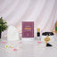 Front display of the Imaan Prestige Men's Hamper arranged on small white pedestals. A maroon Fortress of the Muslim dua book stands in the center, flanked by a white prayer cap, a bottle of attar, and black crystal prayer beads with a gold pendant. The setup is set against a draped white fabric background with warm fairy lights and scattered pink rose petals.