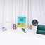Front-facing arrangement of the green-themed kids hamper items, showcasing a folded green plush prayer mat, Basic Duas book, green beaded and digital tasbeehs, crayons, coloring sheet, golden lantern, and butter cookie tin on a white surface with a white curtain backdrop.