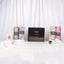A front-facing display featuring a closed black Hadiyah gift box adorned with elegant gold calligraphy and the text "Share the Barakah." Displayed upright on white pedestals around the box are four rolled chiffon hijabs in olive green, purple floral, textured beige, and solid pink. The arrangement also includes a pink velvet box with gold detailing, a beaded pink tasbeeh, a pink digital tally counter, a crystal bakhoor burner, a small jar of bakhoor, and a hijab pin wheel, all set against a soft, draped whi