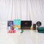 A front-facing arrangement of the green-themed Ultimate Kids Celebration Hamper, displaying a black Hadiyah gift box, a folded green velvet prayer mat, children's Islamic books, crayons, a cookie tin, chocolates, a green tasbeeh, and a mini golden lantern.
