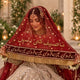A woman in traditional bridal attire wearing a deep red embroidered veil with gold lettering and fringe, over a heavily embellished light-colored gown. She is posed indoors with warm lighting and floral decor in the background.