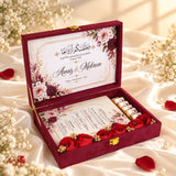 An open burgundy velvet wedding keepsake box with a floral printed insert, containing wedding programs, small wrapped gifts, and red fabric roses, styled on white satin with scattered rose petals and baby's breath flowers.