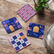 Four geometric patterned coasters on a wooden surface with a plant and glass container in the background.