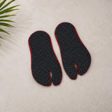 A pair of black tabi-style footbeds with white polka dots and red edges, laid flat on a light beige textured surface with a green leaf in the corner.