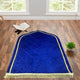 Blue Dome Janamaz with intricate geometric patterns and gold fringe, laid in a bright room with Quran and greenery. Ideal Islamic gift item.