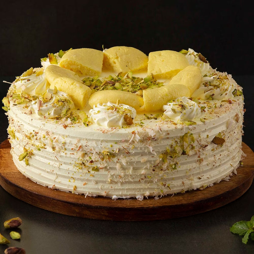 Delicious Rasmalai Cake topped with rasmalai pieces, whipped cream, and crushed pistachios on a wooden base. Buy today for authentic flavors. Main image