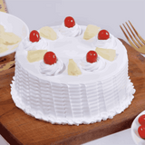 A round pineapple cake with white whipped cream layers and topped with cherries and pineapple segments, presented on a wooden board.
