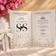 An open white wedding invitation booklet with ornate Arabic and English calligraphy, floral embellishments, and three interlocking ring cutouts in the center. The booklet is styled on a soft white faux-fur surface with pink flowers and a white feather wand in the background.