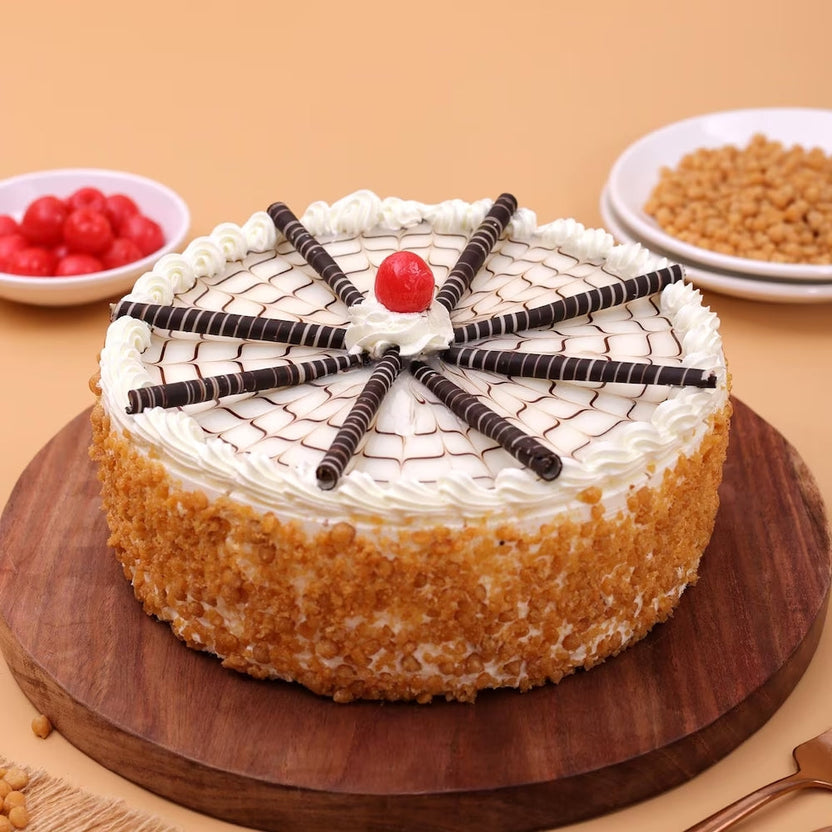 Tantalizing Butterscotch Cake with soft sponge, cream topping, chocolate sticks, and cherry center on wooden board. Order Butterscotch Cake Online now. Main image