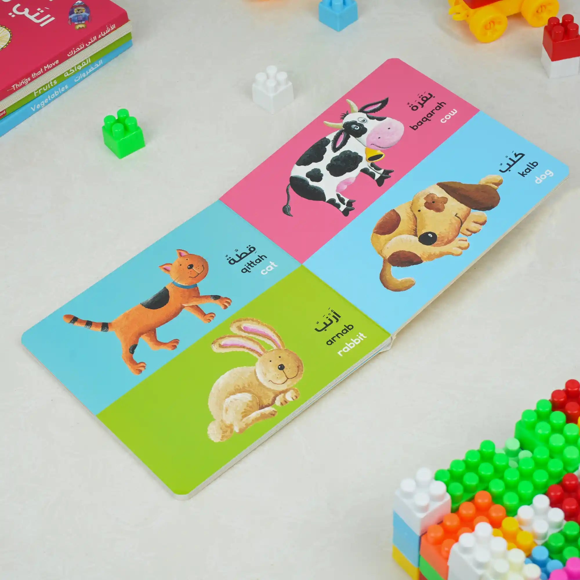 Educational card with animal illustrations and Arabic text on a white surface with toys around.