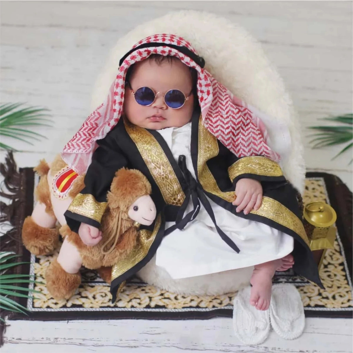 Infant dressed in Baby Boy Bisht Robes with keffiyeh and sunglasses, styled for Eid with plush camel toy and prayer rug.