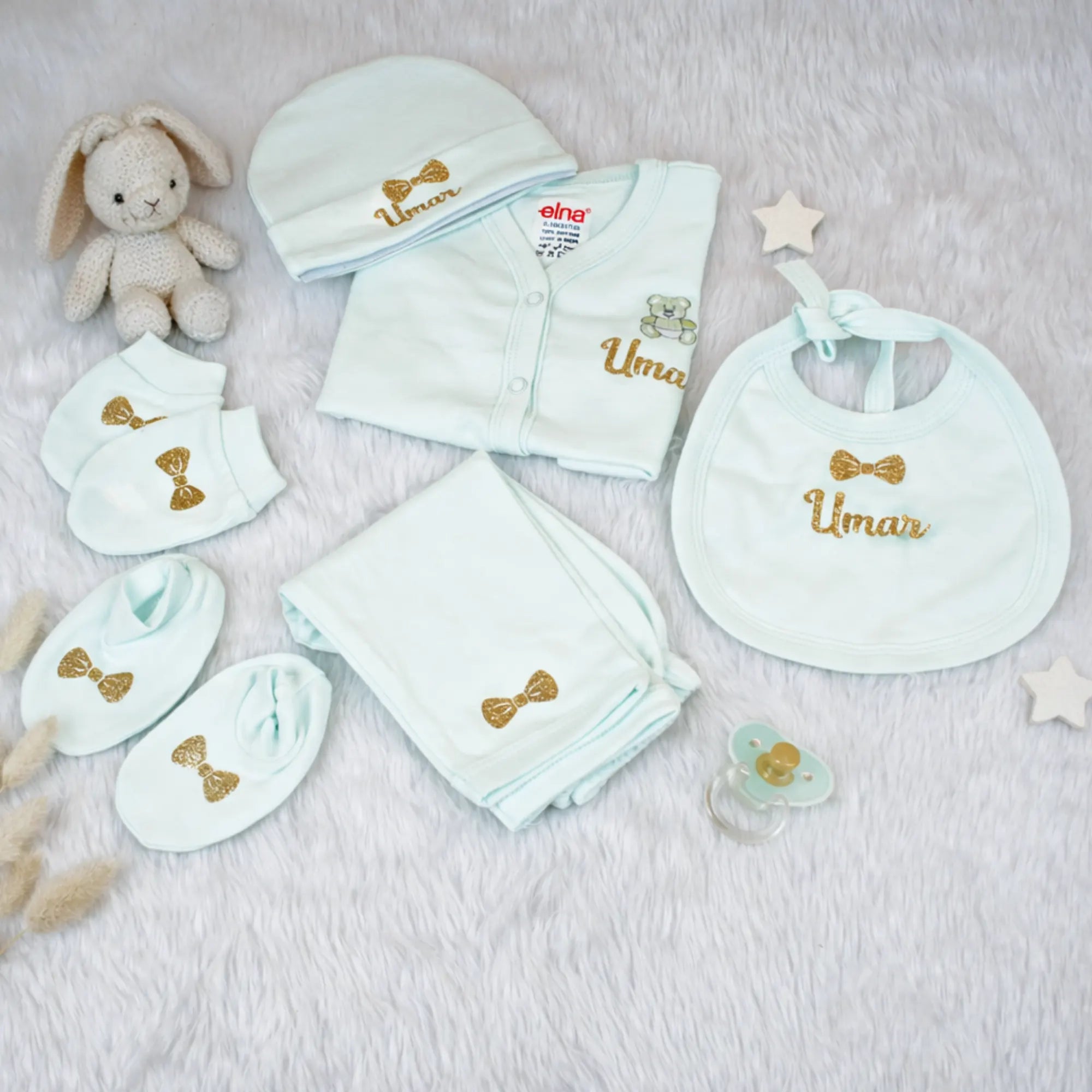 Collection of baby clothing and accessories with 'Umar' embroidered on them, including a onesie, bib, hat, and blanket, on a soft surface.