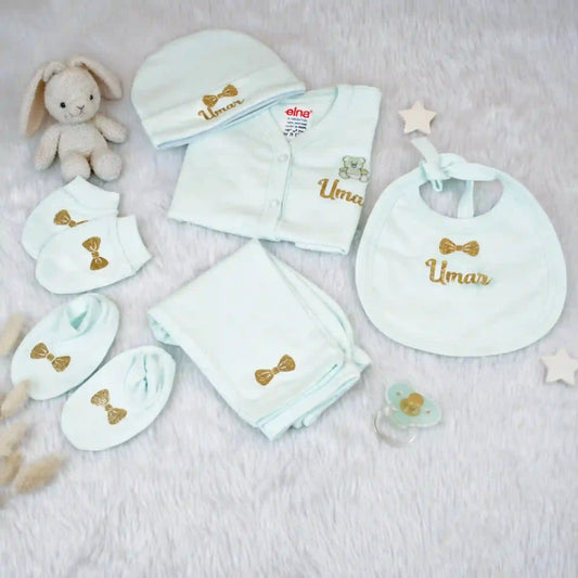 Collection of baby clothing and accessories with 'Umar' embroidered on them, including a onesie, bib, hat, and blanket, on a soft surface.