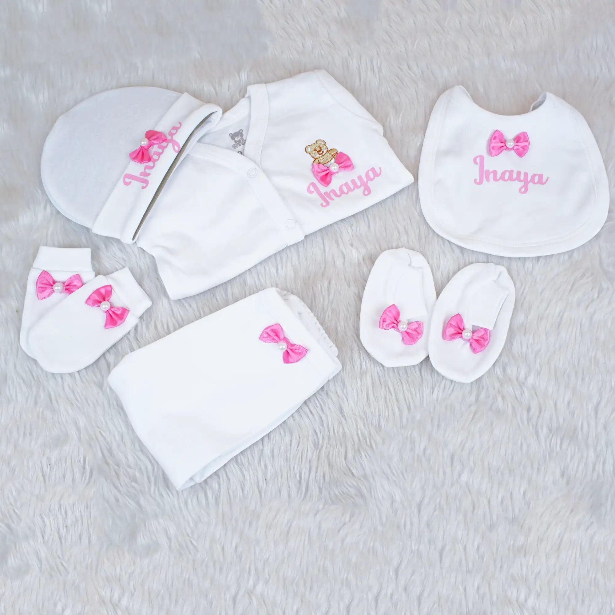 Baby clothing set with white bibs, hats, and socks featuring pink bows and embroidered names on a light gray background.
