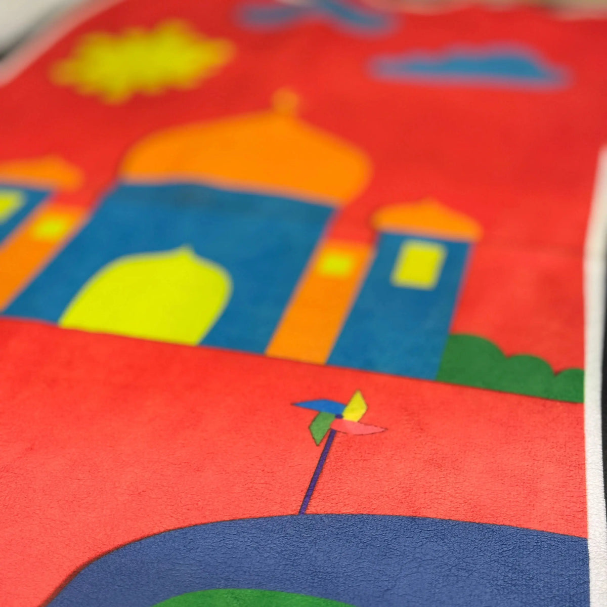 Close-up of Bismillah Adventure Janamaz - Kids Islamic Prayer Mat showing vibrant mosque and road illustrations on soft, foldable fabric.