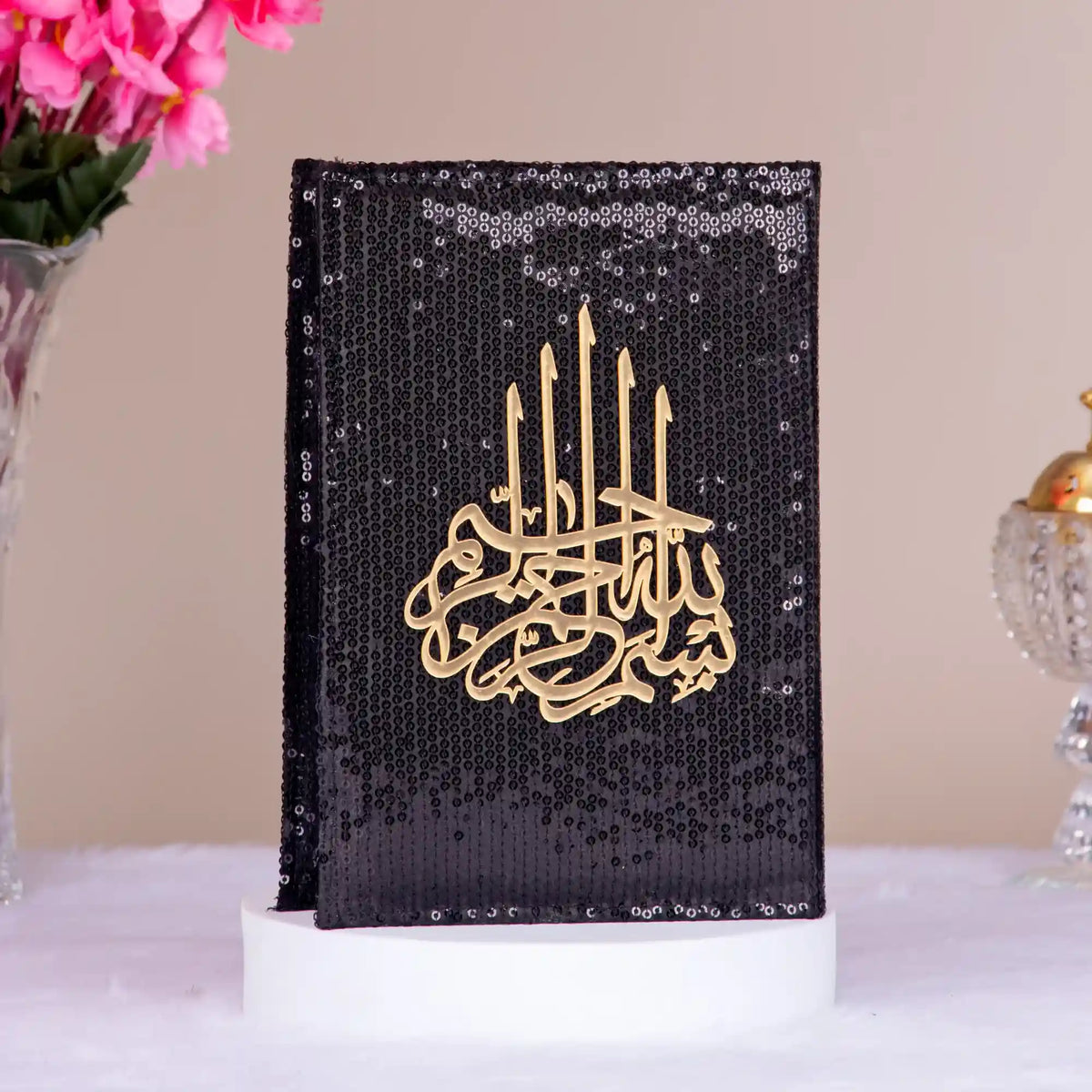Black sequined cover with gold Arabic calligraphy on a white surface with pink flowers and a decorative item in the background.