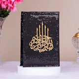 Black sequined cover with gold Arabic calligraphy on a white surface with pink flowers and a decorative item in the background.