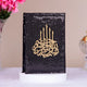 Black sequined cover with gold Arabic calligraphy on a white surface with pink flowers and a decorative item in the background.