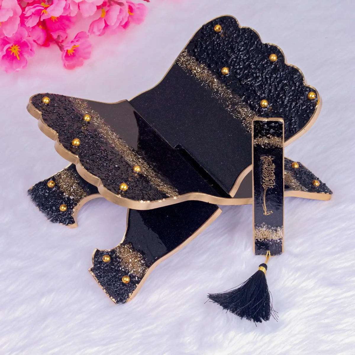 Black and gold decorative item with tassel on a white background with pink flowers.