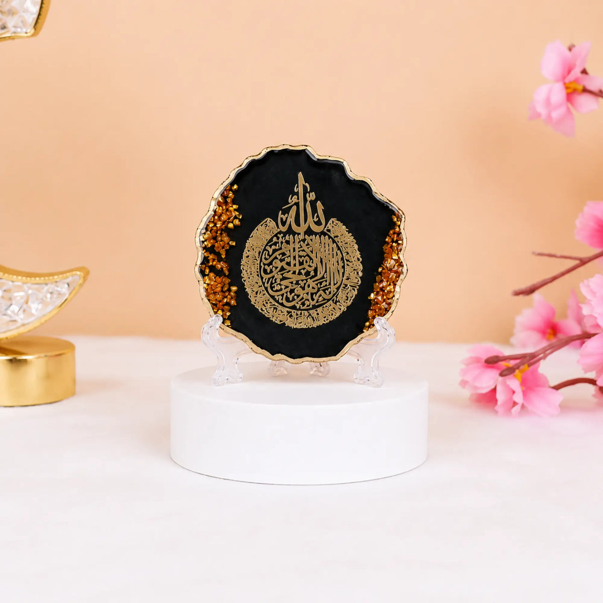 Black 3" Ayatul Kursi Frame with golden flakes, perfect for Ramadan & EID decorations or gifting. Set on a white base with pink flowers nearby.