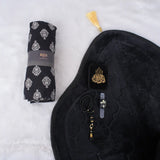 A black-themed Women's Namaz Essentials Hamper flat lay, showcasing a rolled patterned prayer dress, a black velvet prayer mat, a black mini velvet Quran box, black tasbeeh beads, and a digital counter.