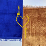 Blue prayer mat with 'Hussain' text and yellow heart, next to a brown prayer mat with intricate patterns.
