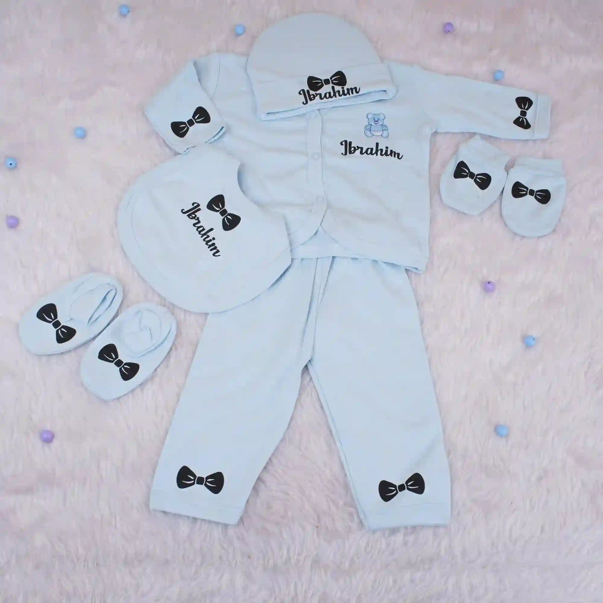 Personalized kids pyjama set in light blue with custom name 'Ibrahim', includes cap, mittens, bib, and pants with bow prints, ideal for newborns.