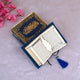 Open Blue Stones Mini Tilawat-e-Quran showing Arabic script pages with gold feather bookmark, placed beside a designer velvet box.