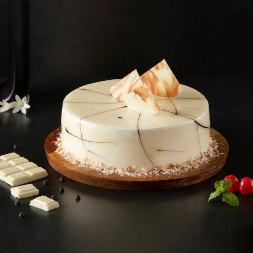 Buy Hazelnut Chocolate Cake Online - Nutty Richness Delight featuring a glossy white chocolate glaze, chocolate swirl design, and nut garnish on a wooden base. Main image