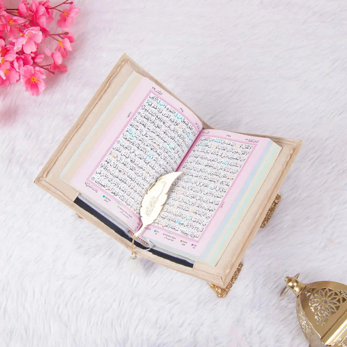 Open book with decorative cover on a white surface, surrounded by pink flowers and a small lamp.