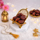 A crescent-shaped golden acrylic date stand holding a cluster of dates, beautifully displayed on a white hexagonal base amidst festive Ramadan decor, including a small lit gold lantern, a pearl tasbih, gold star ornaments, and pink flowers on a soft white faux-fur surface.