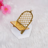 A close-up of a single, empty crescent-shaped gold acrylic date stand resting on a white hexagonal block, set against a soft white fluffy background with blurred pink flowers.