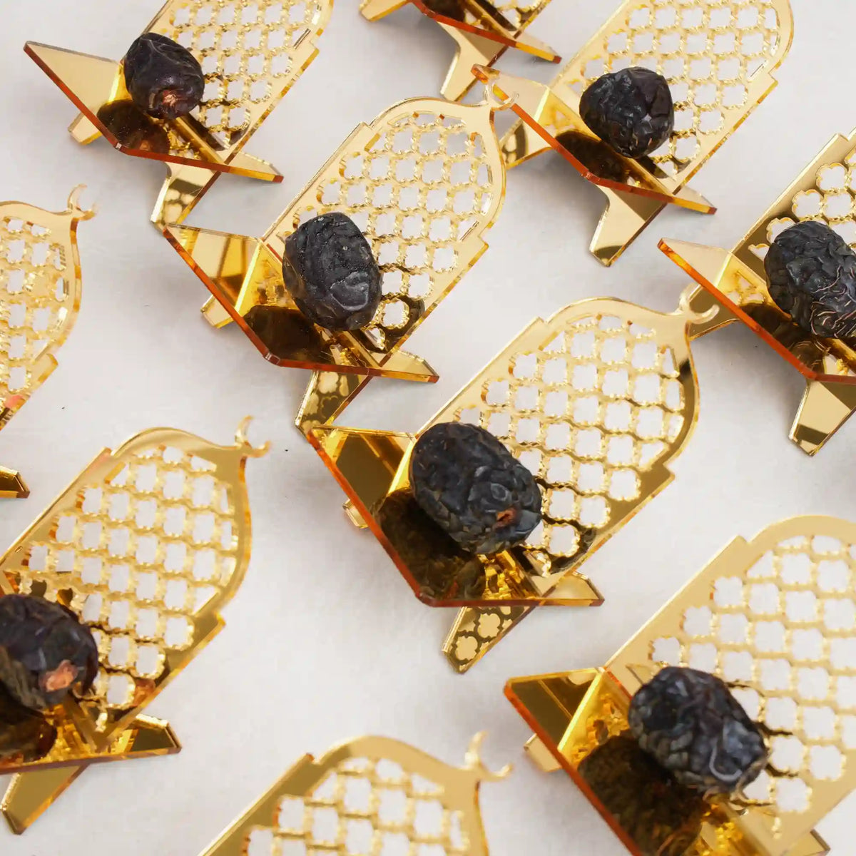 An overhead view of multiple crescent-shaped gold acrylic date stands arranged in rows on a white surface, with each golden stand holding a single dark date.
