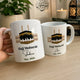 Two mugs with Hajj Mubarak designs on a wooden surface, with a hand holding one mug.
