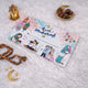 A single beautifully designed white Eid Mubarak money envelope displayed on a fluffy white surface, elegantly styled with wooden prayer beads, golden crescent moon props, and a small decorative lantern.