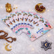 A fanned-out stack of white Eid Mubarak Eidi envelopes featuring colorful cartoon illustrations of Muslim families celebrating, placed on a white faux fur surface alongside a bowl of dates and a golden lantern.